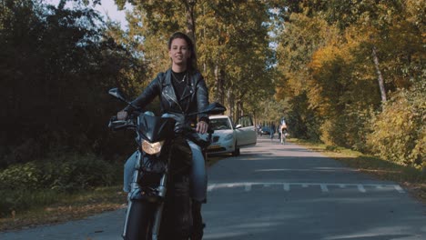 Hübsche-Lächelnde-Europäische-Junge-Frau,-Die-An-Einem-Sonnigen-Tag-Ein-Motorrad-Trägt,-Das-Lederjacke-Im-Wald-Mit-Lebhaften,-Bunten-Goldenen-Herbstblättern-Trägt