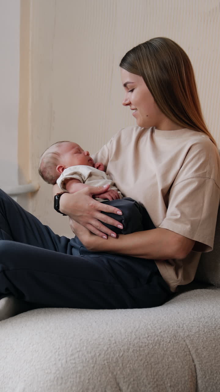 Mommy waving her lovely newborn in hands. Woman sits on sofa holding her sleeping baby and looking at him with a smile.. Vertical video
