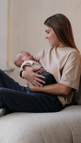 mommy waving her lovely newborn in hands. woman sits on sofa holding her sleeping baby and looking at him with a smile.. vertical video