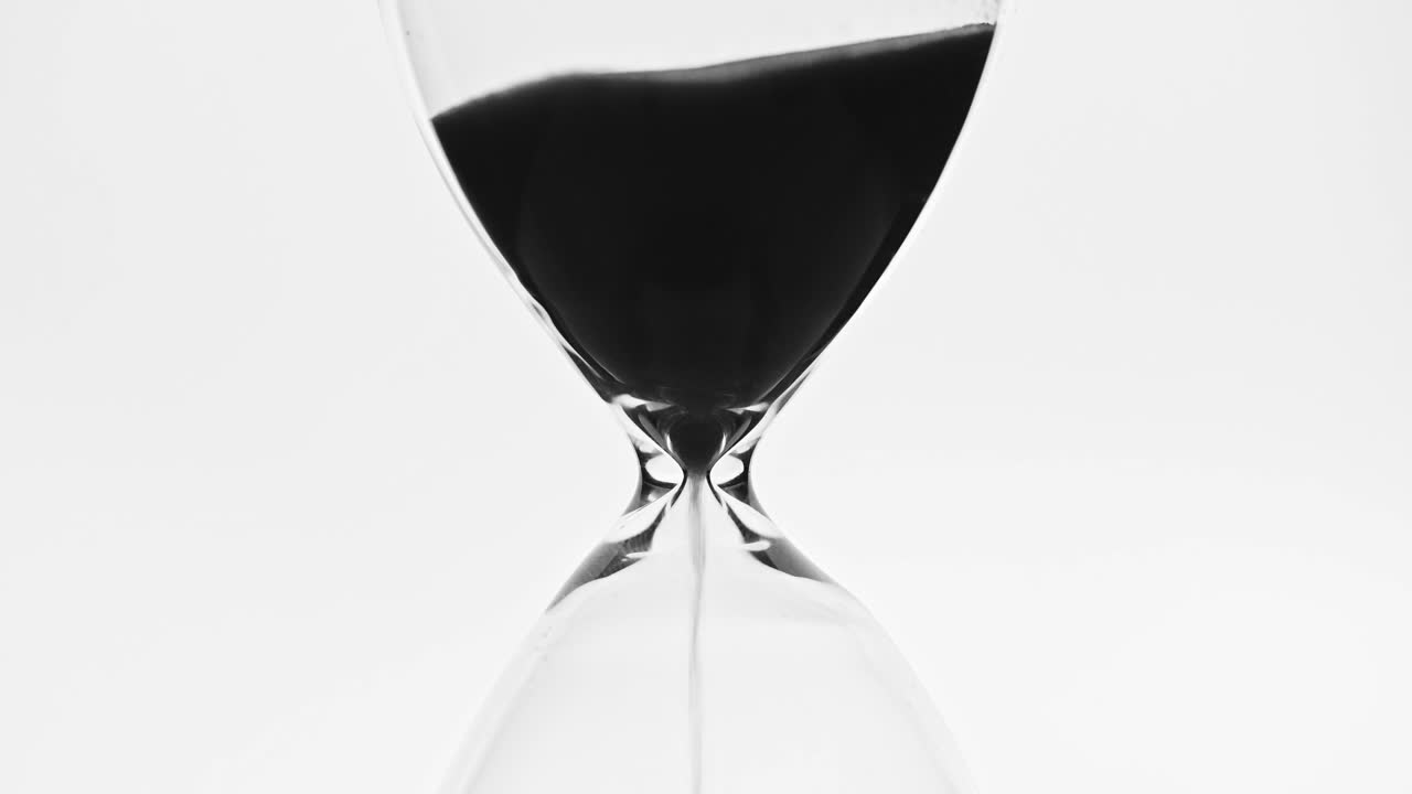 A close-up video of an hourglass with black sand steadily flowing from the upper chamber to the lower one. The minimalist monochrome composition symbolizes the passage of time.