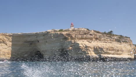 Alfanzina-Lighthouse,-Algarve,-Portugal-atop-cliffs-with-caves-from-a-speeding-speedboat