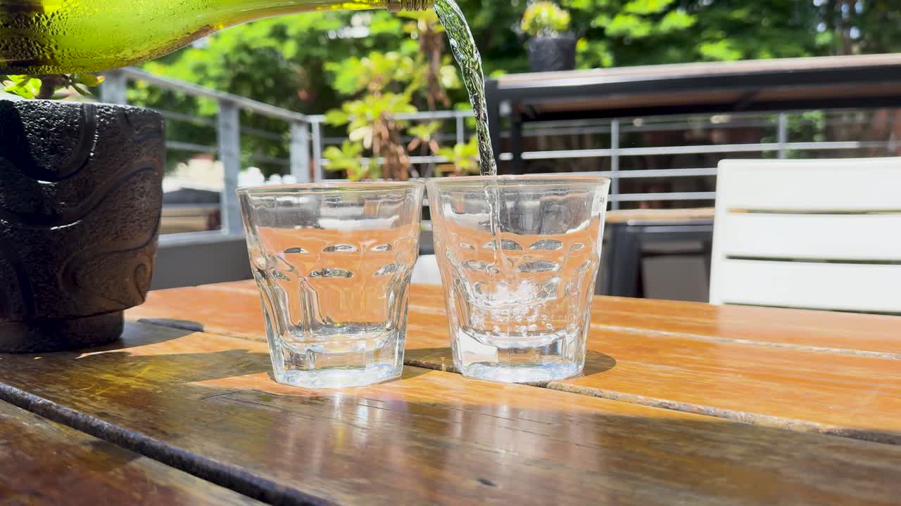 A bottle pours water into glasses on a sunlit outdoor table, creating a refreshing scene with vibrant greenery