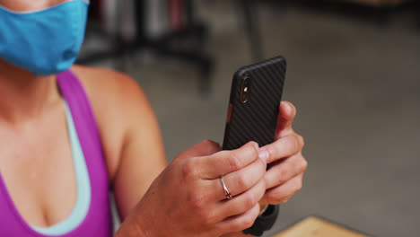 Caucasian-woman-wearing-face-mask-using-smartphone-at-cafe-in-gym