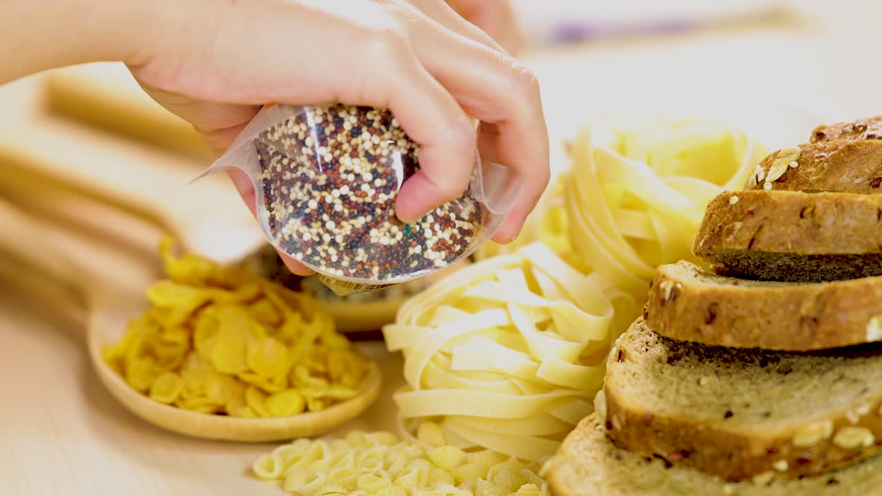 A hand pours quinoa onto a platter with sliced bread, pasta, and cornflakes in a well-lit kitchen setting