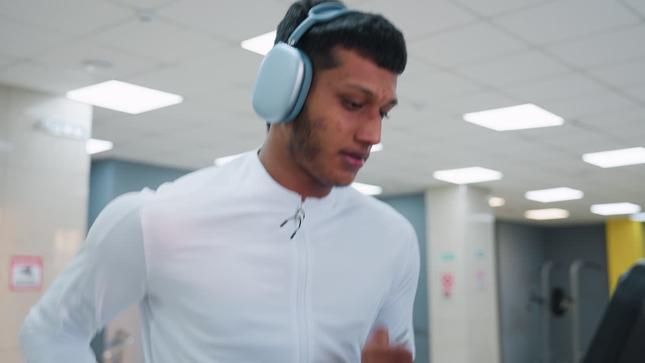 young man in white athletic top runs on treadmill in bright modern gym, wearing headphones, focused on workout with blurred equipment and urban view through large windows