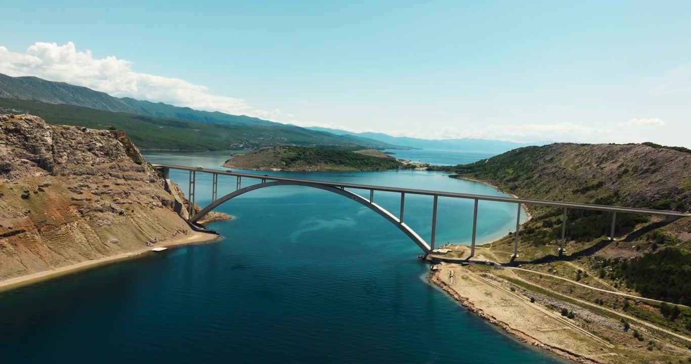 Krk Bridge crossing turquoise Adriatic water between rocky coastal hills in Croatia. Mountains, islands, shoreline, and blue summer sky fill the distance.