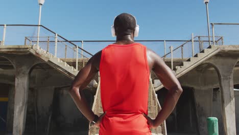 vista traseira de um homem afro-americano com fones de ouvido, de pé, fazendo uma pausa no exercício ao ar livre