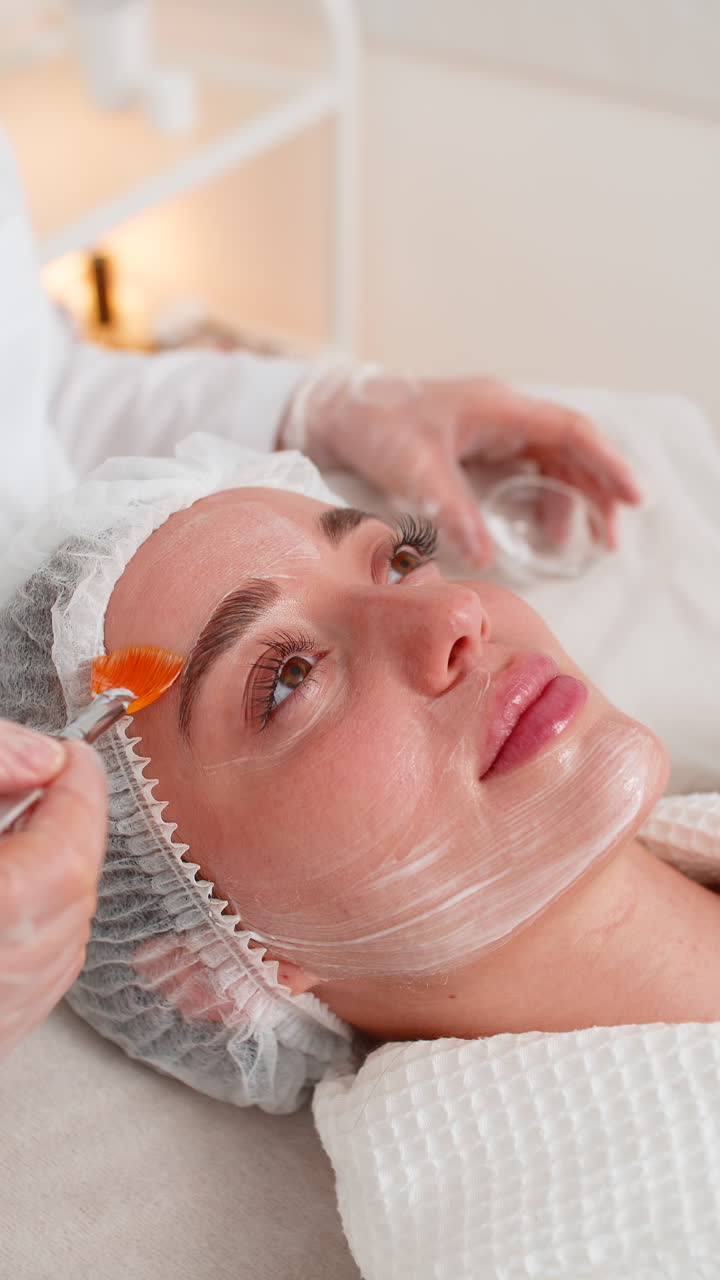 Estetista che applica una crema medica idratante sul viso di una giovane donna con dei pennelli durante una terapia enzimatica