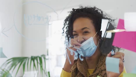 Woman-wearing-face-mask-writing-on-glass-board-on-her-desk-at-office