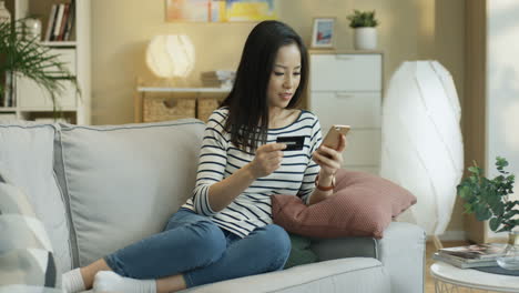 young charming woman sitting on the sofa in the living room with a smartphone and credit card in hands and shopping online