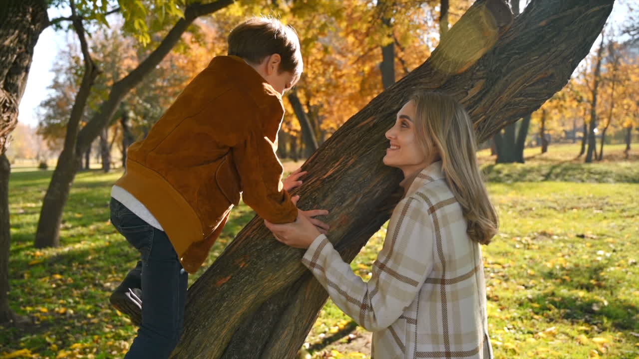 Happy family in an autumn park. Mother holds her son while he is climbing on a tree. Slow motion