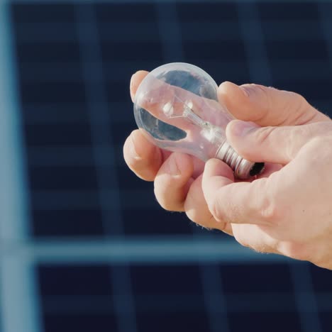 Side-View-Of-A-Man's-Hands-Holding-A-Light-Bulb-Against-The-Background-Of-Solar-Panels