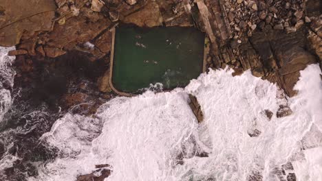 aerial top-down descending shot of rock pool with swimmers in winter morning – ocean waves, foam, cloudy sky, bird’s eye view