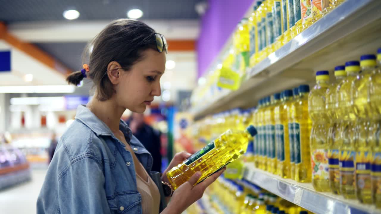 chica en la tienda, leyendo la pegatina de información en una botella de aceite. seleccionado una botela de aceite de girasol, lo pone en el carrito. variedad de productos en la fila