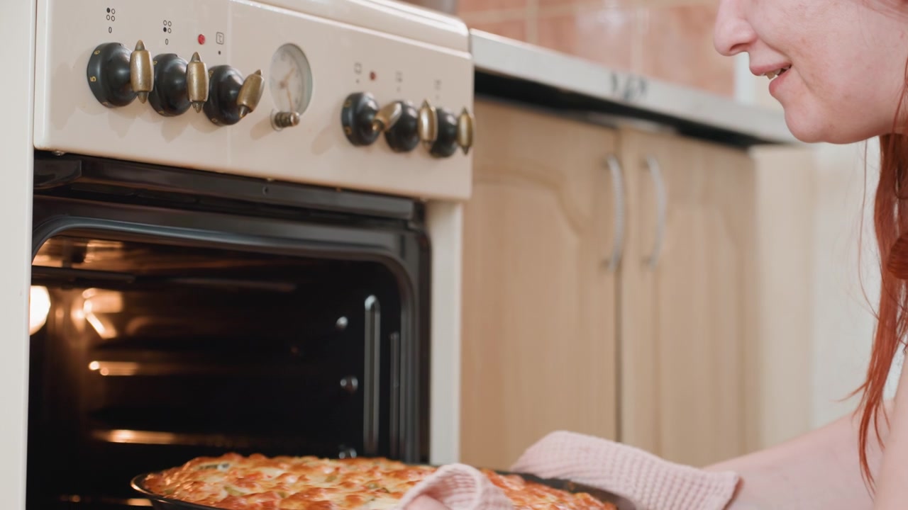 Primo piano di mani che indossano guanti testurizzati che estraggono una teglia dorata da un forno a incasso, mostrando l'attenta rimozione del piatto caldo, un movimento costante verso l'esterno, luce calda della cucina e cucina casalinga sicura
