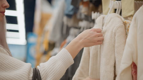 Stylish woman with white hair hangs back child hoodie sweater on rack in store, inspecting plush texture and hood detail against blurred pastel garments and modern ambient lighting