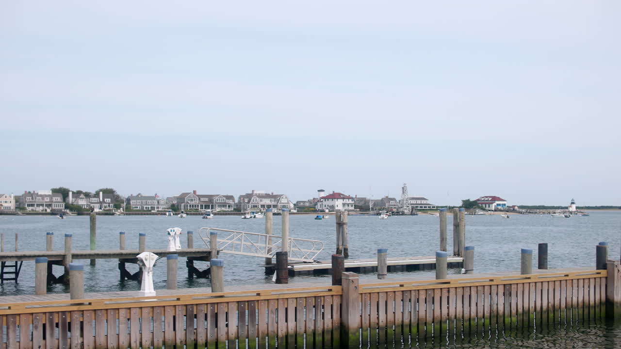 Jævn panorering til højre langs Nantucket Havn, Massachusetts, der afslører kajpæle, rolige bugtvande og klassiske New England-huse ved vandet under en blød himmel