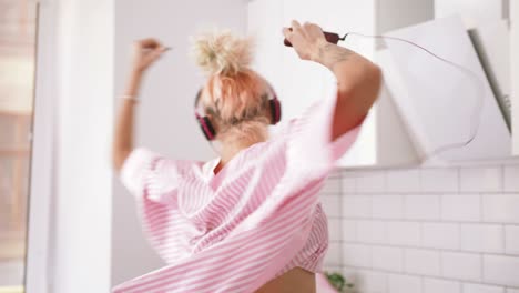 happy young woman with pink hair dancing in kitchen wearing pink pajamas and listening to music with headphones