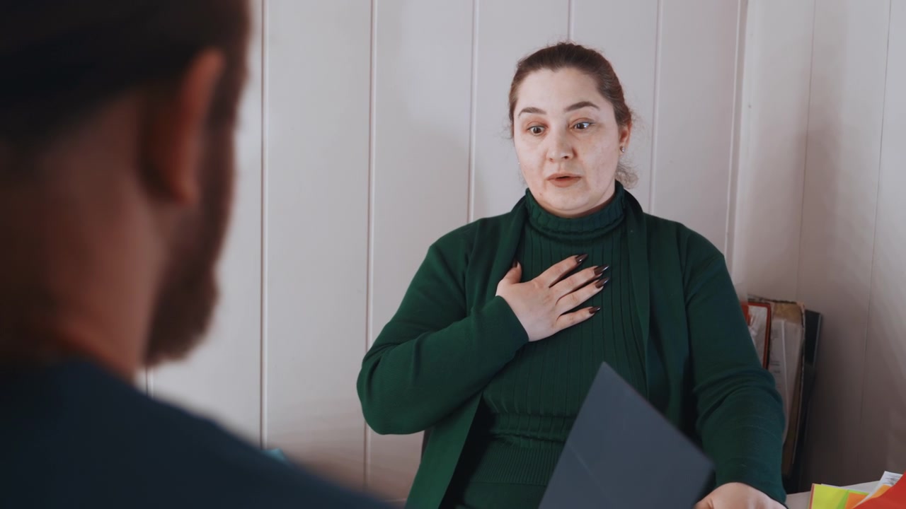Ev ortamında göğüs rahatsızlığı olan bir kadını değerlendiren paramedik, görevli panoyu tutarken hasta elini göğsüne koyuyor, sakin profesyonel diyalog, klinik jestler ve dinleme, sıcak ev ortamı.