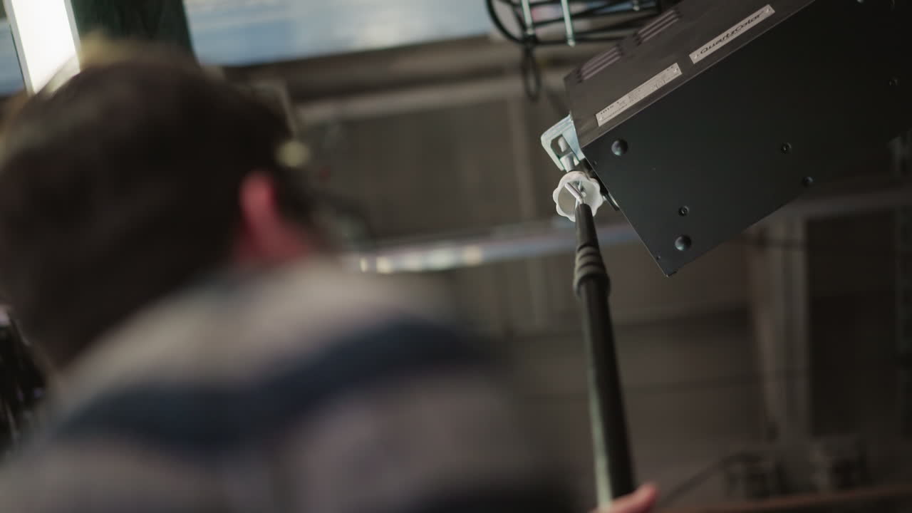 Technician using boom pole to adjust lighting equipment in broadcast studio. Professional media production environment with visible equipment and studio setup in background