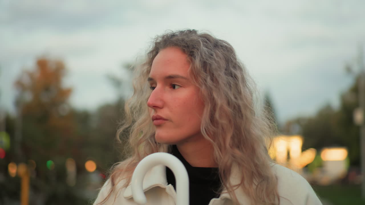 Calm lady walking looking unhappy gazing around holding white umbrella with blurred bokeh background filled with lights trees city elements under soft sky during peaceful solitary stroll
