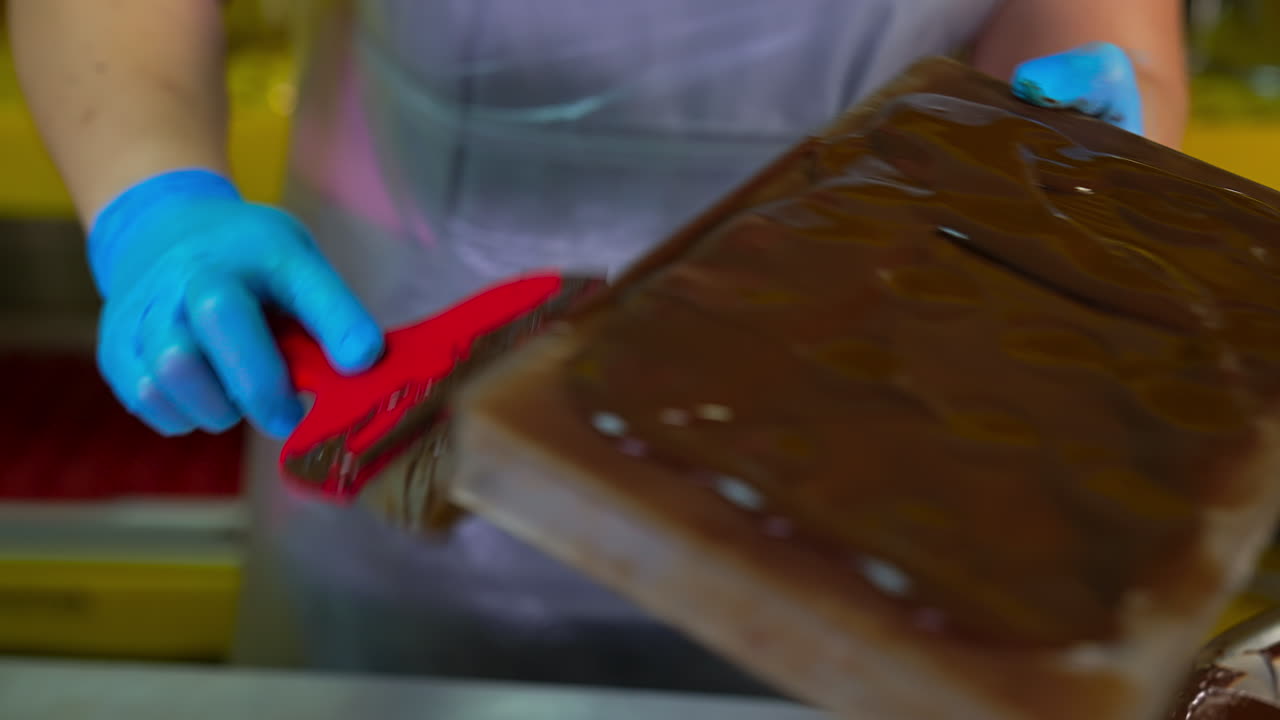 Chocolatier covers the filled molds with final layer of chocolate. Hands in blue gloves take off the spare chocolate from the molds with the help of spatula. Close up.