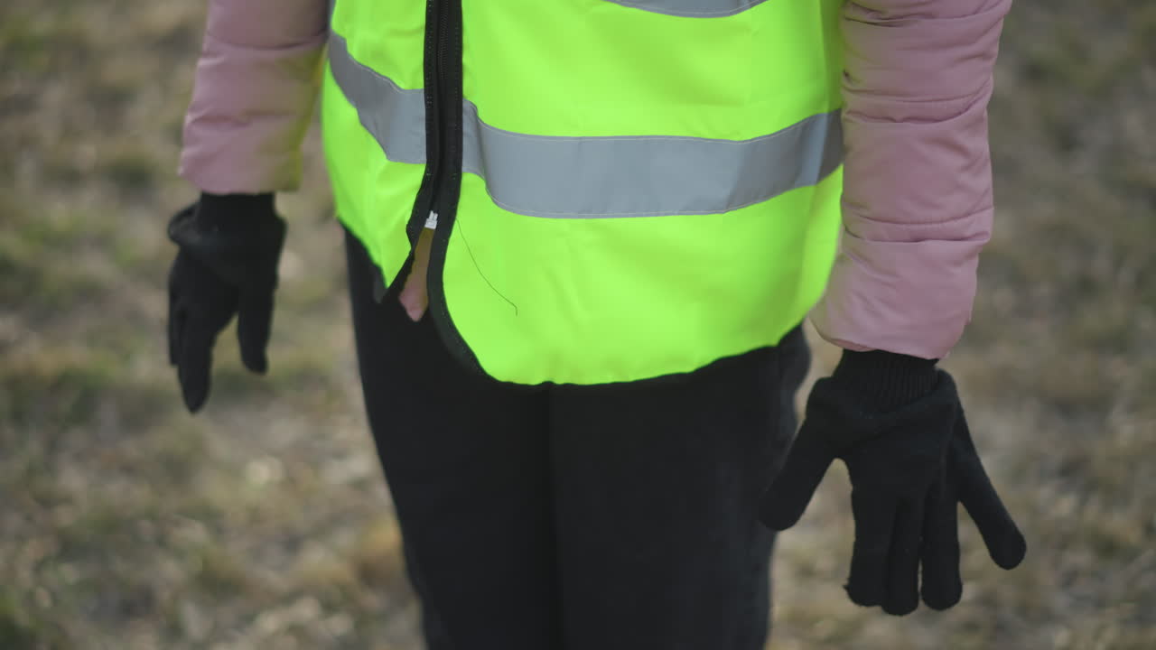 Person wearing neon yellow reflective safety vest over pink padded jacket putting on black gloves outdoors in cold weather, preparing for outdoor work or activity in grassy environment during winter