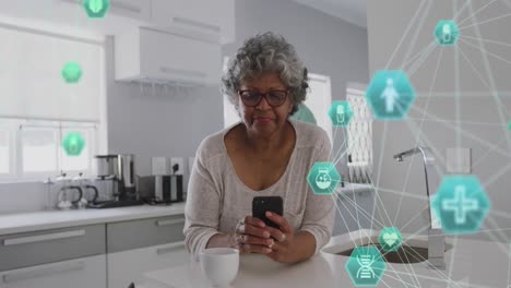 animation of connected icons forming globes, senior african american woman using laptop in kitchen