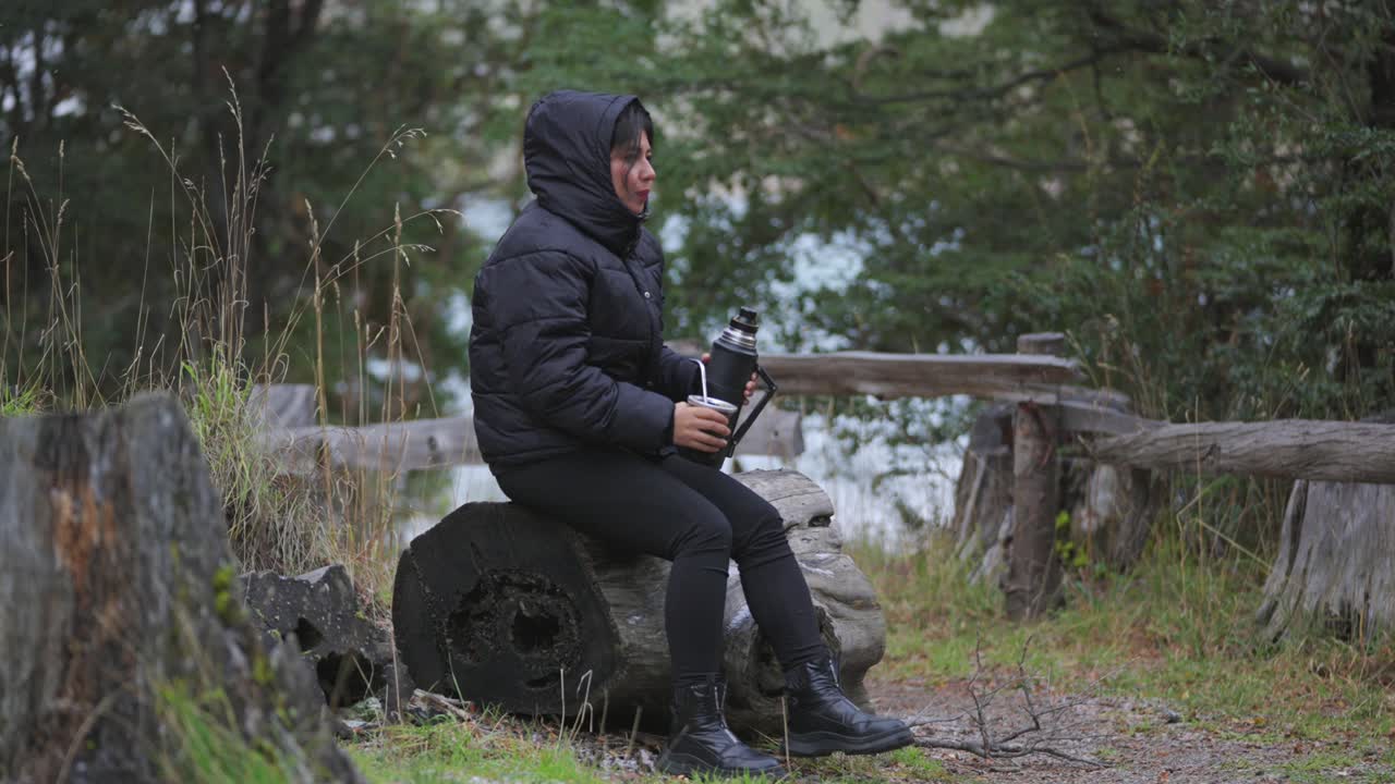 Woman seated on log, drinking Yerba Mate amid snowy grass and pine trees of Bariloche, Argentina