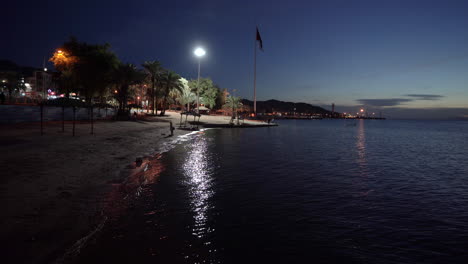 Ein-Licht-Spiegelt-Sich-Auf-Der-Wasseroberfläche-Im-Golf-Von-Aqaba-Mit-Einem-Riesigen-Fahnenmast-Im-Hintergrund