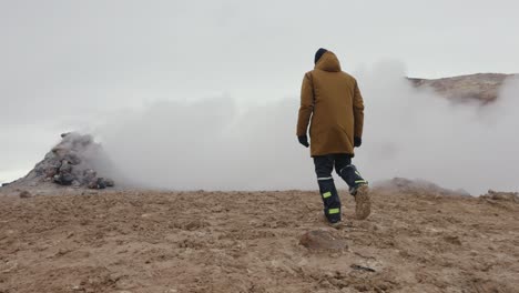 tourist slowly walk on orange mud toward dense white vapor cloud, iceland