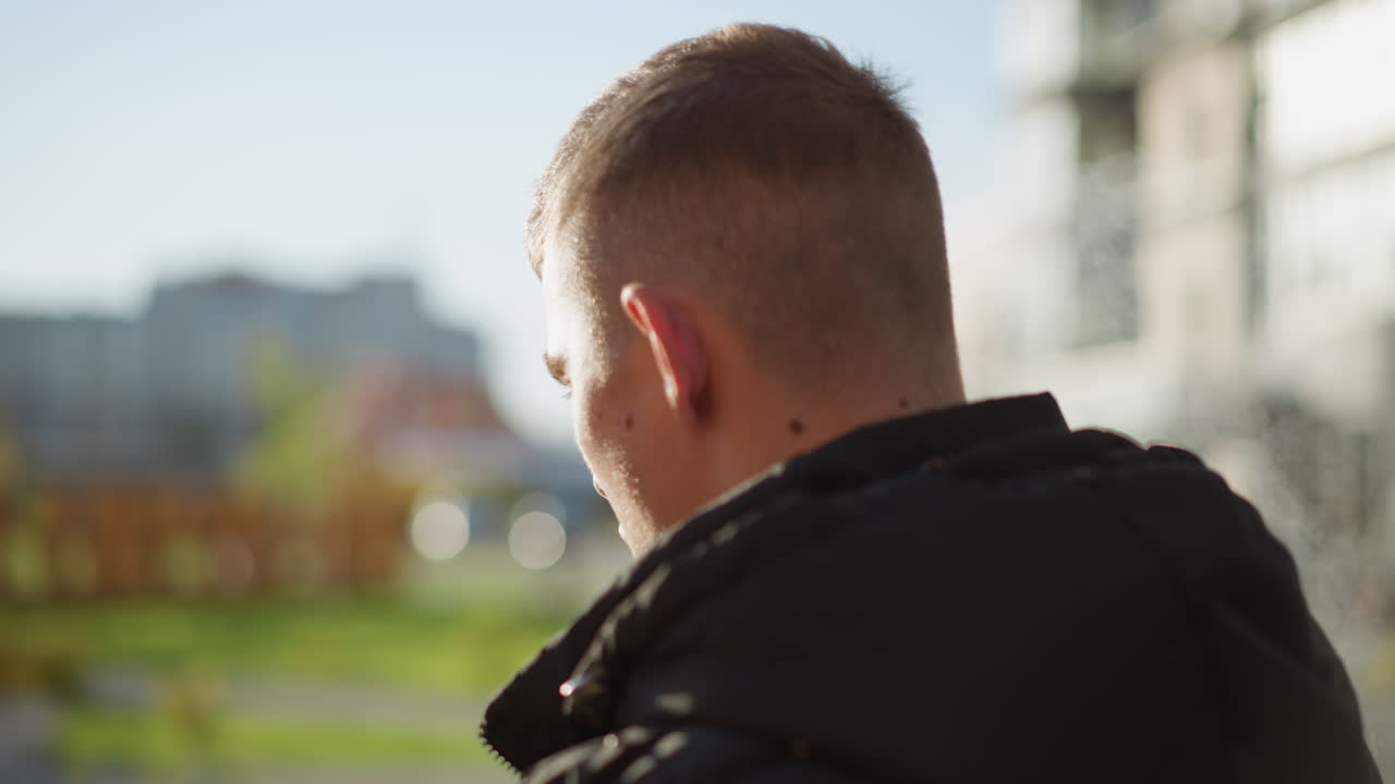 back view young man exhales shisha smoke outdoors with sunlight softly illuminating side of face in vibrant urban setting with blurred background buildings and greenery in natural afternoon light
