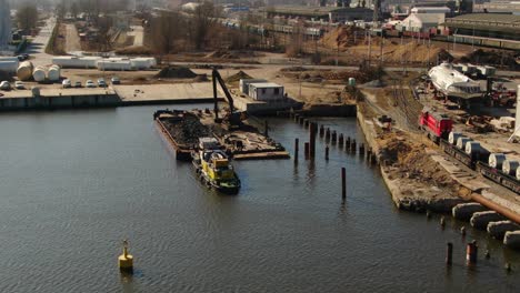 drone shot of a excavator dumping rubble from the boat into water near a port shore