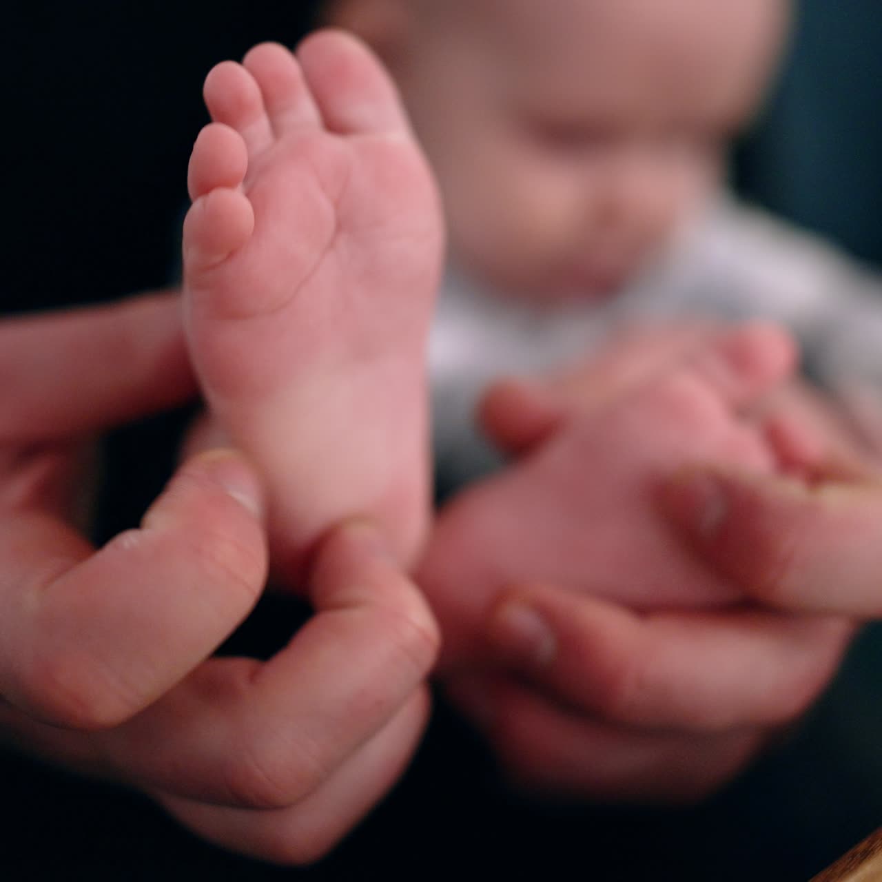 Dad's hands hold the tiny feet of his baby. Father tickles and claps son's feet. Close up. Blurred backdrop