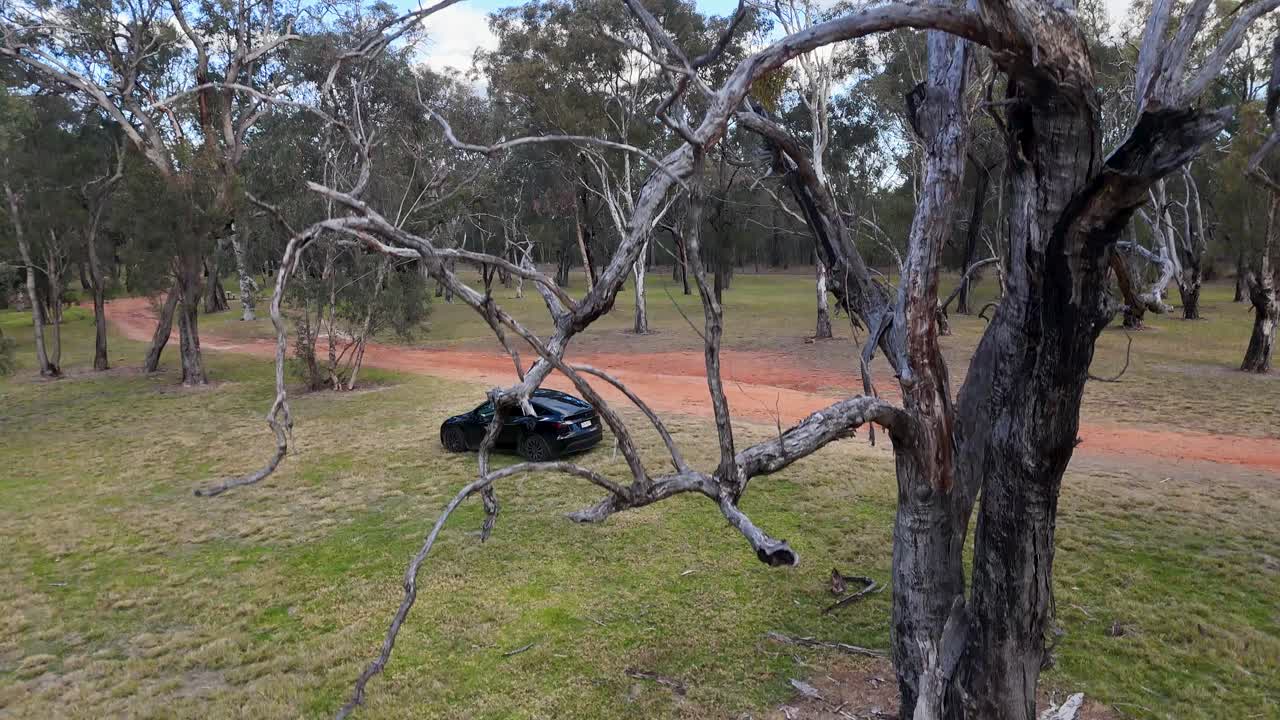 A black electric vehicle travels along a winding dirt road in a grassy, wooded park under daylight, captured by a smooth aerial camera movement