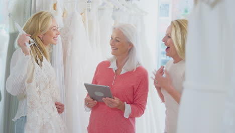 Sales-Assistant-With-Mother-Helping-Adult-Daughter-To-Choose-Wedding-Dress-In-Bridal-Store