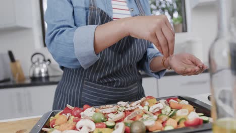 Midsection-of-biracial-woman-seasoning-chopped-vegetables-on-baking-tray-in-kitchen,-slow-motion