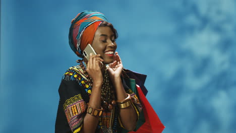 joven afroamericana con turbante y ropa tradicional hablando por teléfono y sosteniendo bolsas de compras