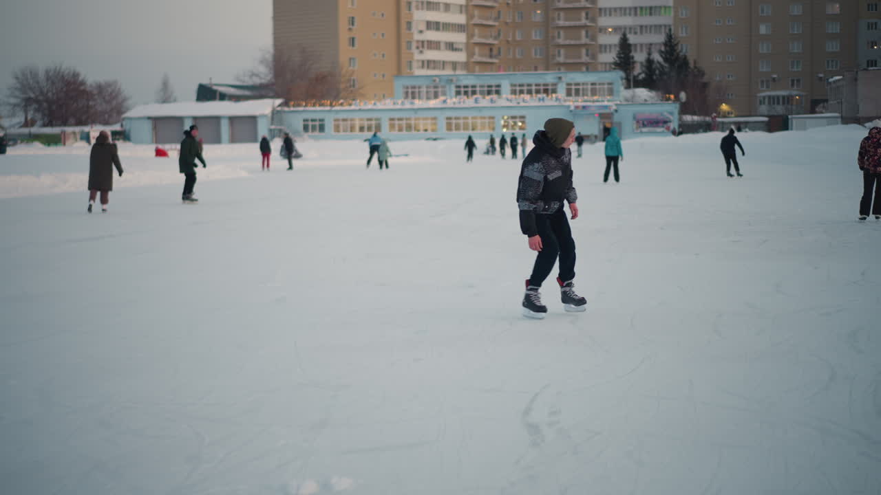 expert skater showcasing advanced moves on snowy urban ice rink, smooth gliding and confident posture against backdrop of fellow skaters and residential buildings under soft winter light