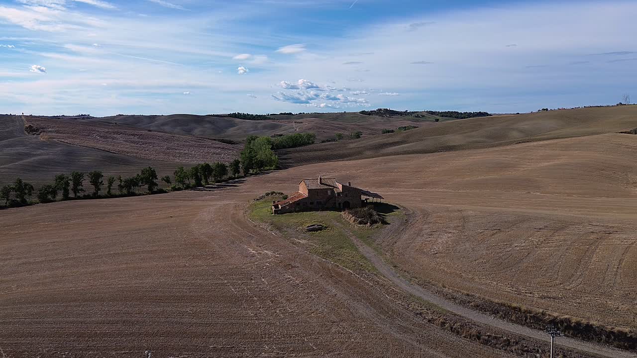 Rustic farmhouse surrounded by rolling hills, dry fields and serene rural landscape in Tuscany under a blue sky.