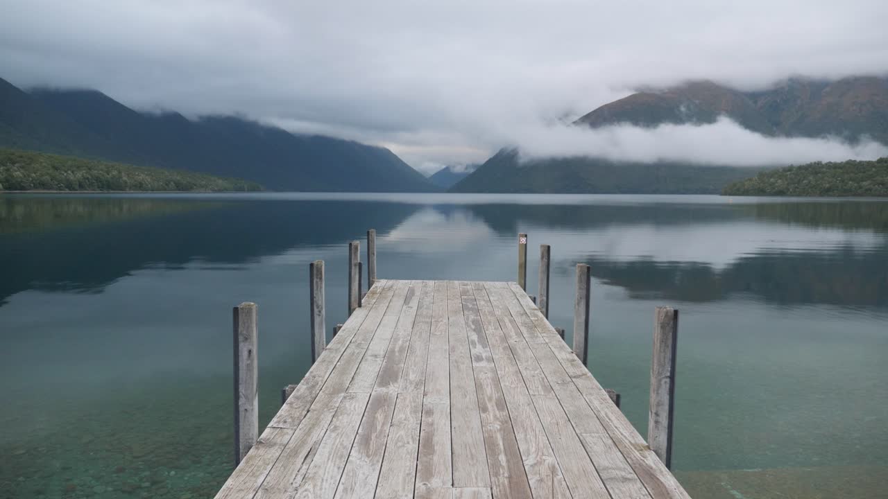 Static shot of the iconic Rotoiti lake wooden jetty New Zealand on a cloudy early morning in 4K with mountains reflection in crystal clear water.