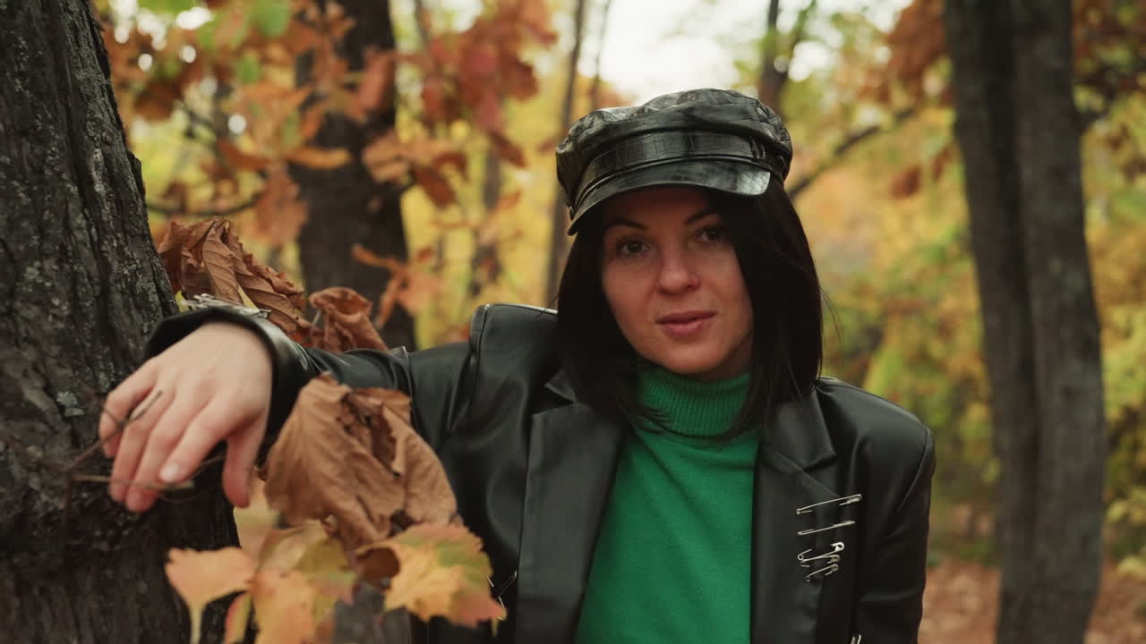 woman in leather jacket leaning on tree in autumn woods, confident rocker pose, studded blazer, green turtleneck, direct gaze, hands resting on bark, warm foliage backdrop, fashion editorial mood