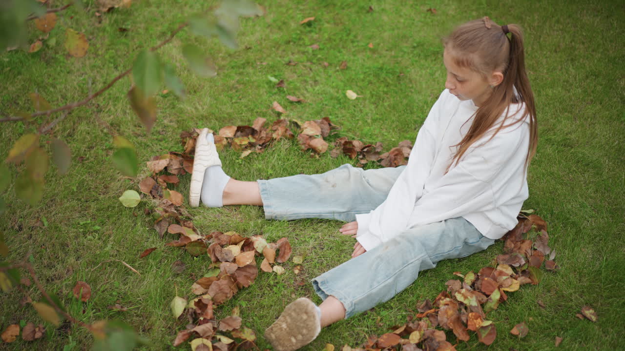 Girl sitting in leaf circle on grass, white sweater and jeans, gazing thoughtfully with legs outstretched, scattered brown leaves forming ring, soft overcast light, quiet backyard atmosphere, gentle