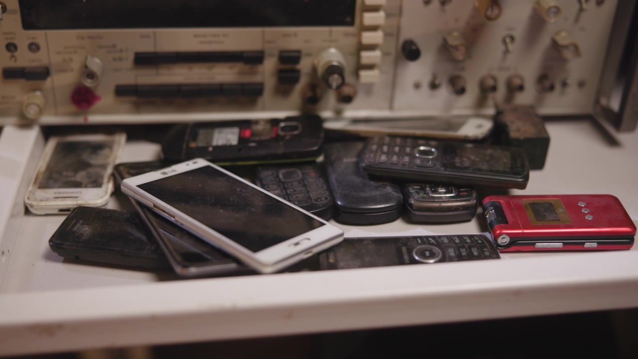 Low-angle view of used mobile phones, remotes, and handheld devices stacked on workshop bench beside control panel, with worn surfaces, cables, tools, muted light, and shallow focus