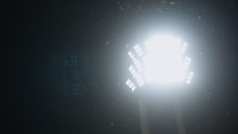 powerful stadium light beams through falling snowflakes in cold night sky, creating dramatic atmospheric glow with scattered particles illuminated midair and subtle silhouettes in background