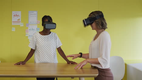 two diverse female coworkers standing at desk, testing vr googles