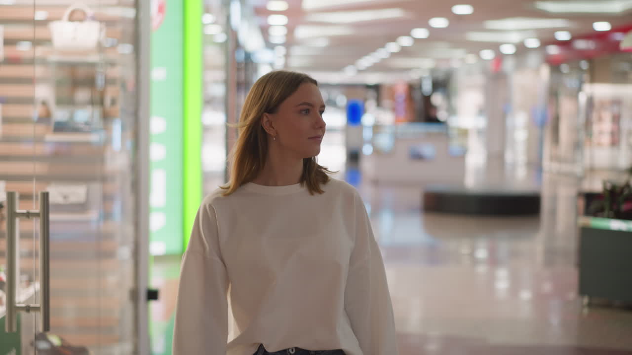 Shopper in white top walking through shopping mall, glancing around with colorful lights and blurred glass background, capturing a casual and modern retail environment