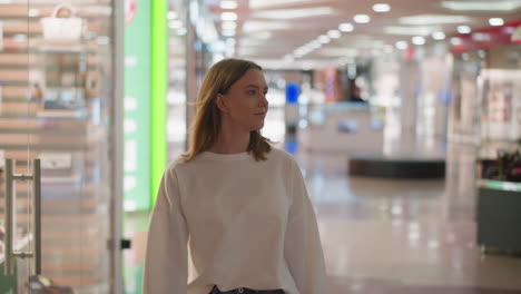 shopper in white top walking through shopping mall, glancing around with colorful lights and blurred glass background, capturing a casual and modern retail environment