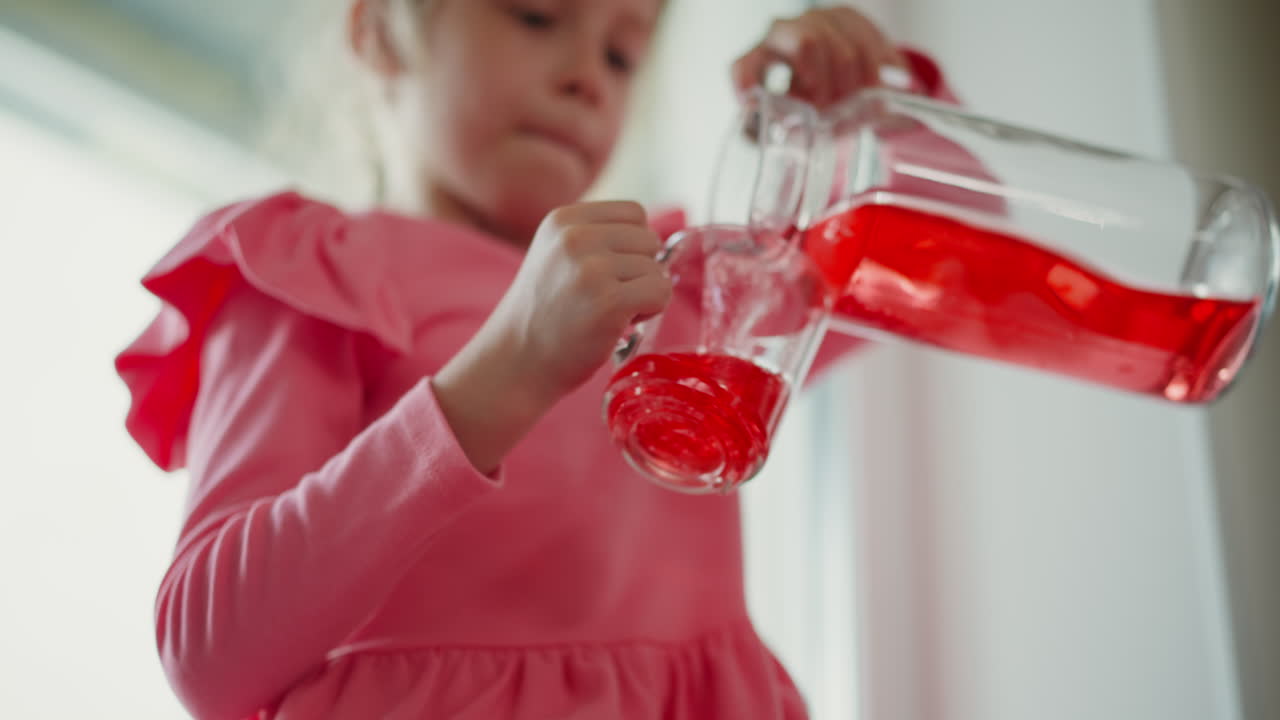 Bright eyed girl carefully pouring vibrant red juice from glass jar into clear glass, small hands gripping handle, sunlight illuminating flowing liquid and focused expression on young child