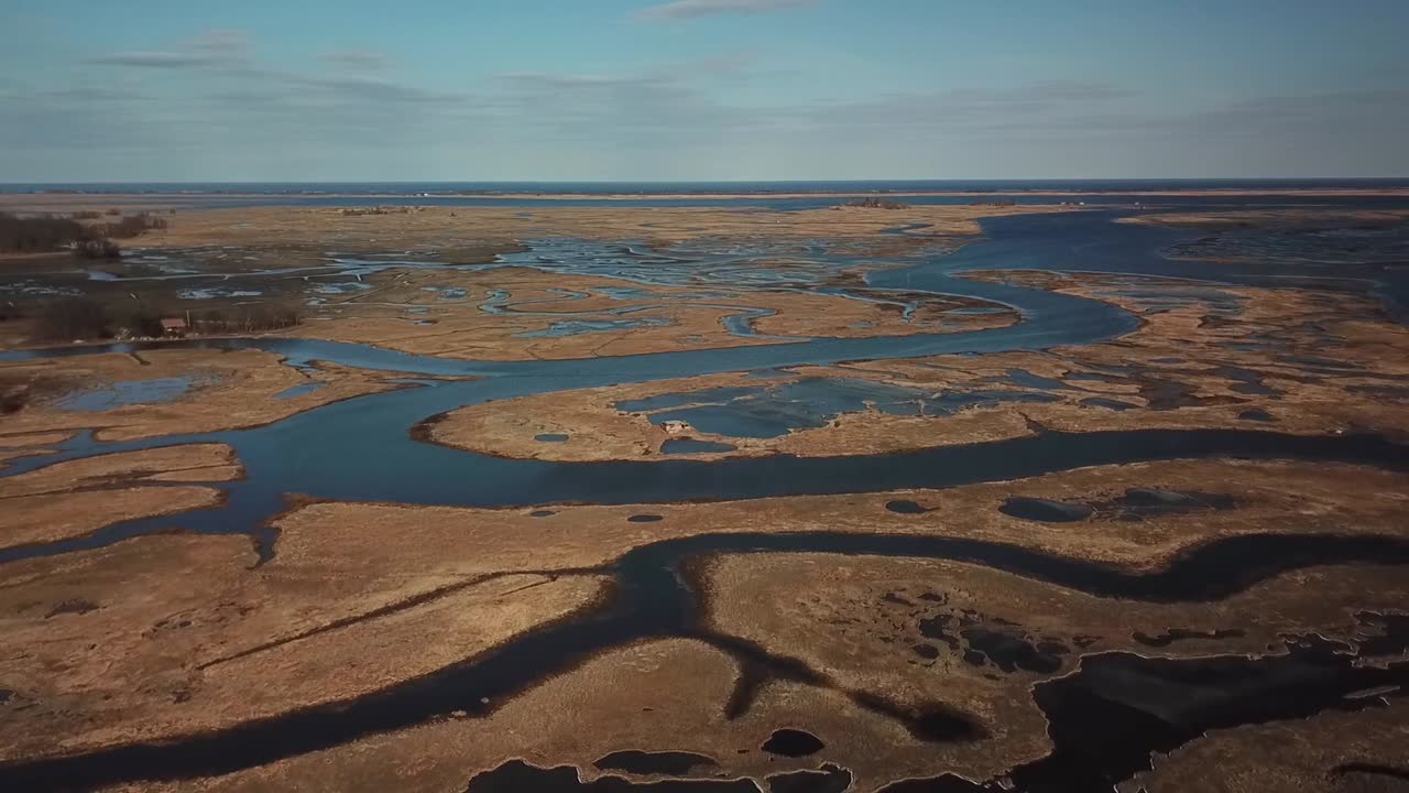 Bred droneoptagelse af Essex Bay og Great Marsh, Massachusetts, med bugtende tidevandskanaler, der skærer gennem gylden marsk mod Atlanterhavet i klart dagslys.